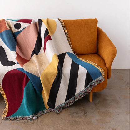 A multicolor abstract patterned throw blanket with fringed edges draped over an orange armchair in a minimalist room setting.