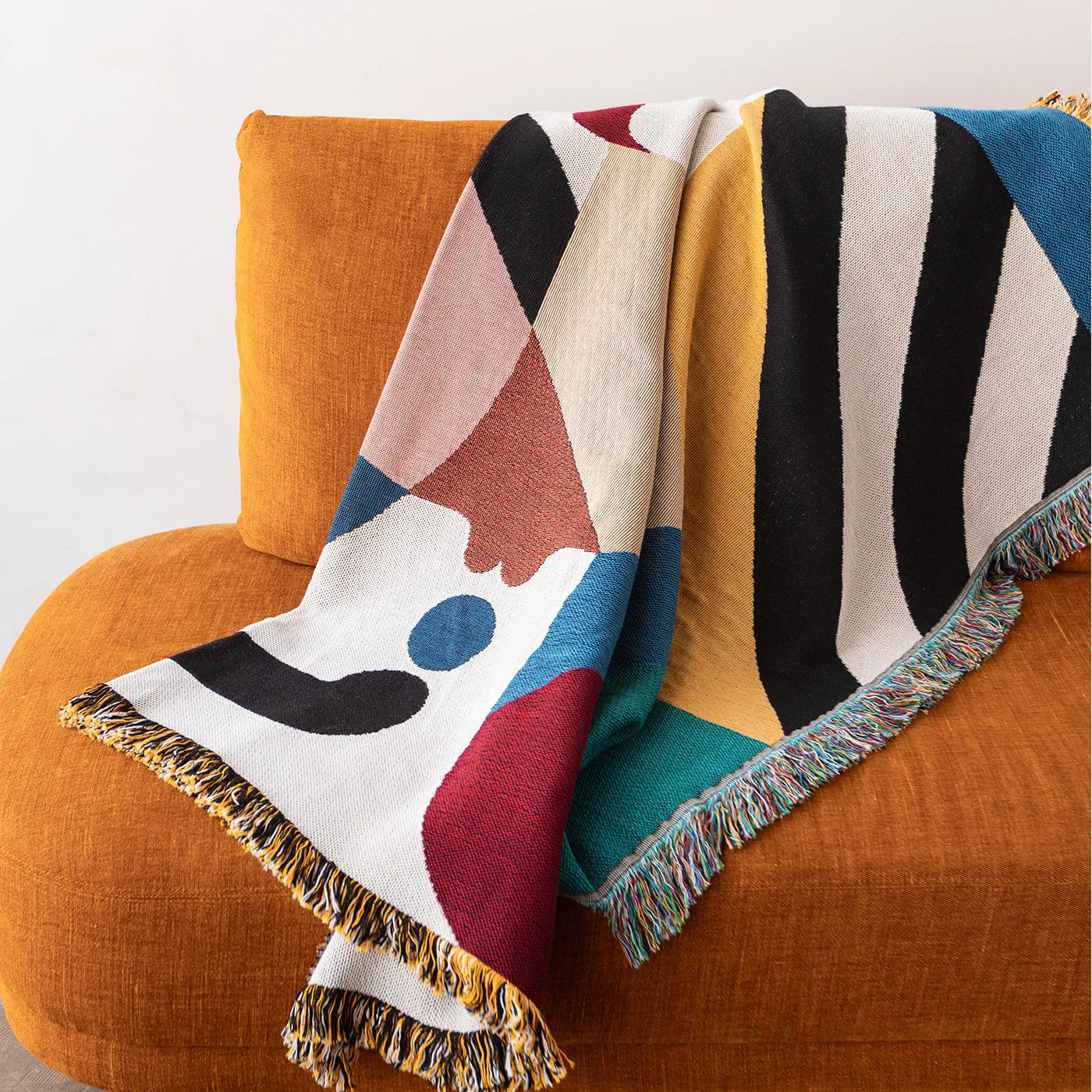 A multicolor abstract patterned throw blanket with fringed edges draped over an orange armchair against a white wall.