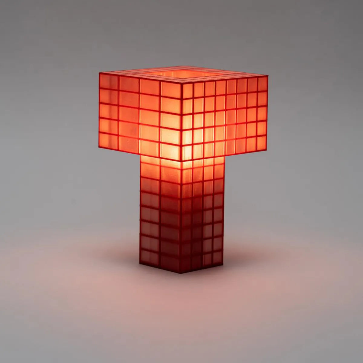 Illuminated Ridezign Tesser 5:8 table lamp in Cherry with an open top, made from a red grid over warm orange panels, casting a soft glow against a neutral grey-brown background.