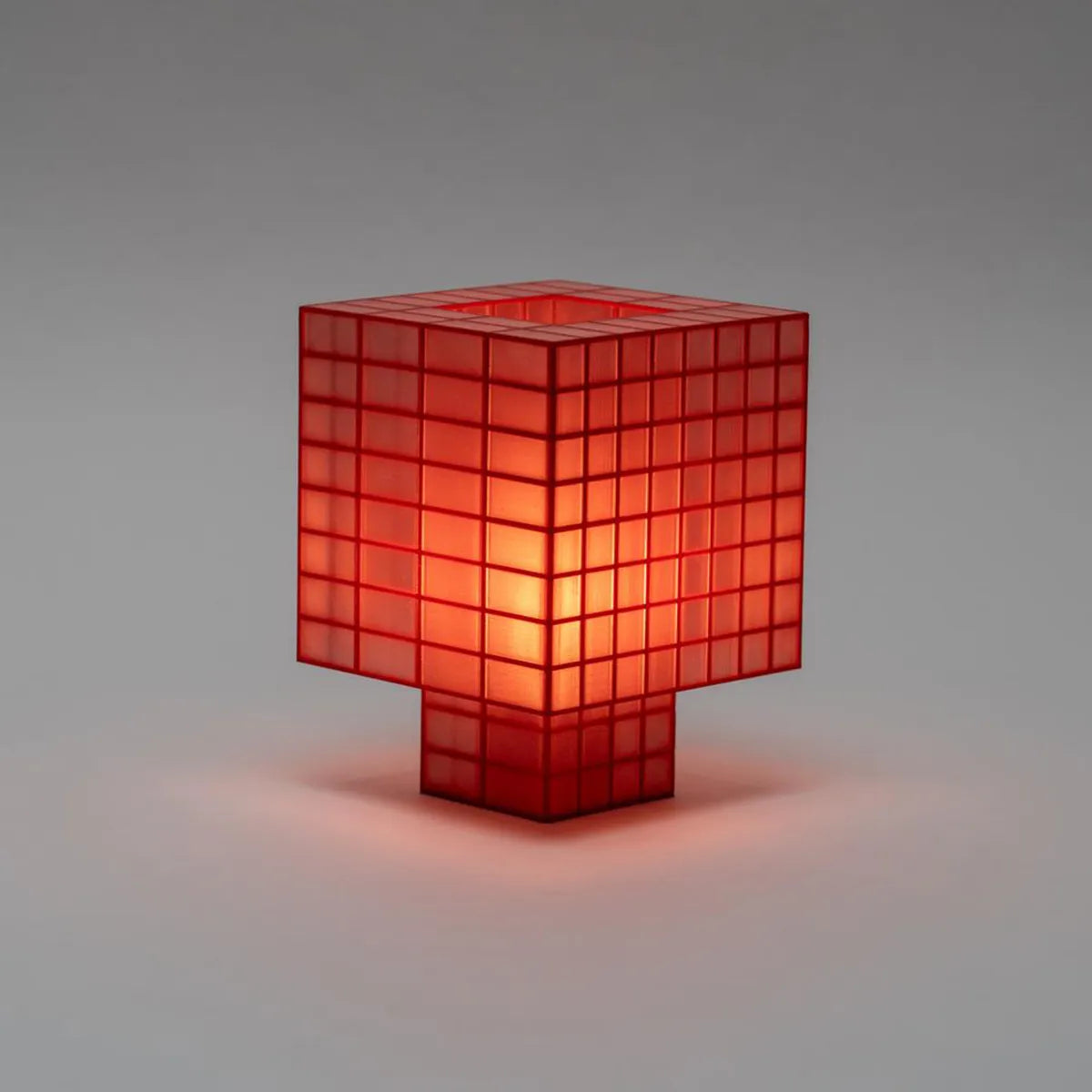 Illuminated Ridezign Tesser 8:3 table lamp in Cherry with an open top, made from a red grid over warm orange panels, casting a soft glow against a neutral grey-brown background.