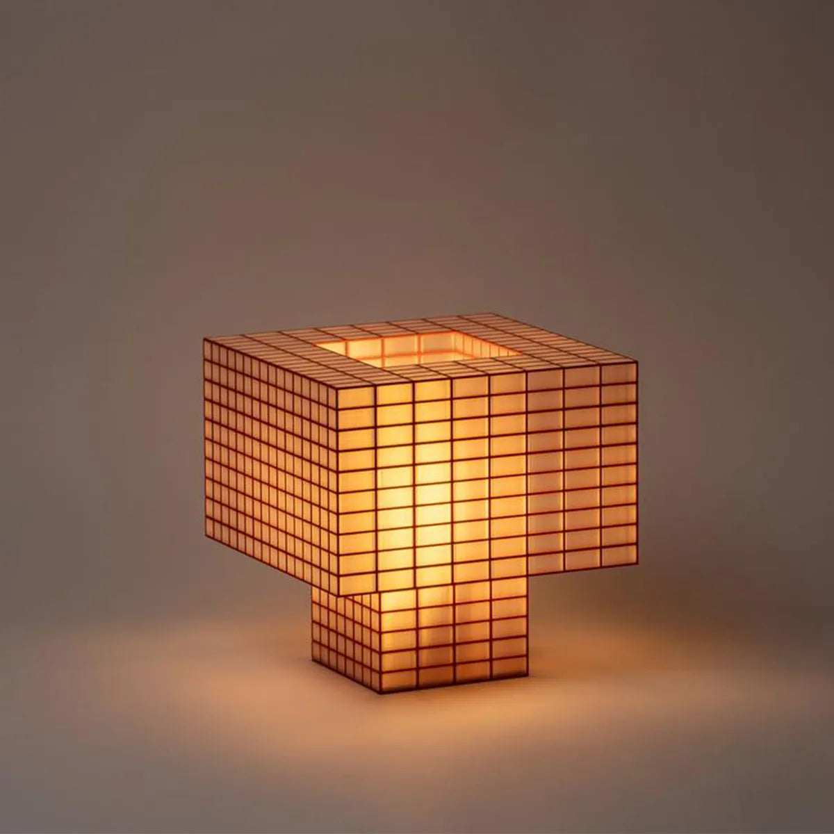 Illuminated Ridezign Tesser XL 5:3 lamp in Granite with an open top, made from a brick red grid over warm yellow panels, casting a soft glow against a neutral grey-brown background.