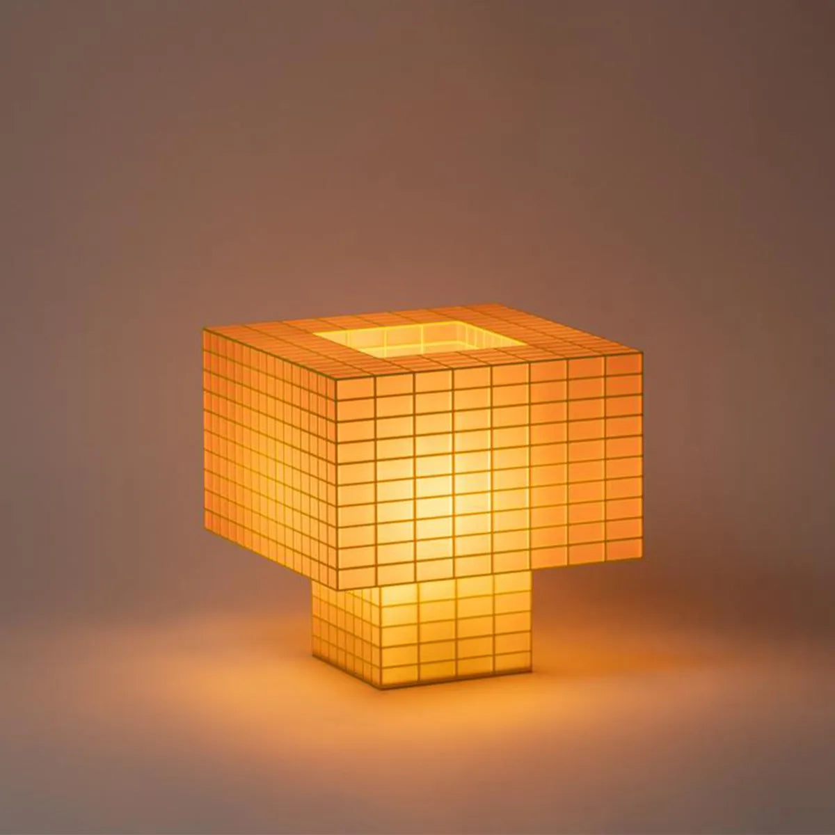 Illuminated Ridezign Tesser XL 5:3 lamp in Guava with an open top, made from a yellow grid over warm orange panels, casting a soft glow against a neutral grey-brown background.