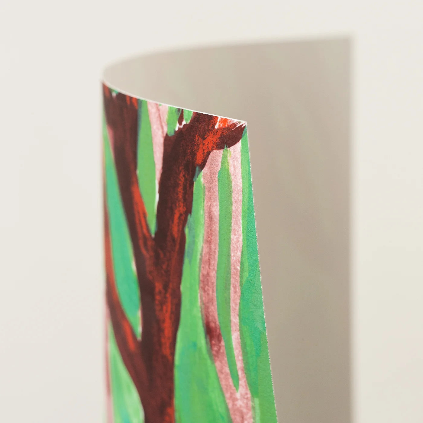 A close-up of an art print showing a watercolor tree design with green background, reddish-brown branches, and pale pink vertical streaks, photographed against a light neutral backdrop.