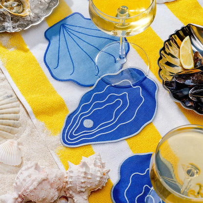 Table setting with blue shell coasters, white wine glasses, and seashells on a yellow and white tablecloth.
