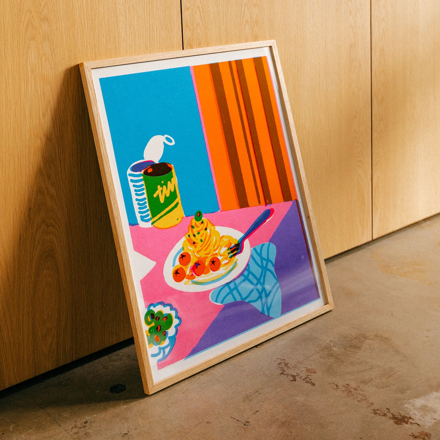A framed colorful art print leaning against a light wood cabinet on a concrete floor. The wall art features a stylized still life with a plate of spaghetti and tomatoes, a green striped can, a blue chair, and bold blocks of blue, orange, pink, purple, and yellow.