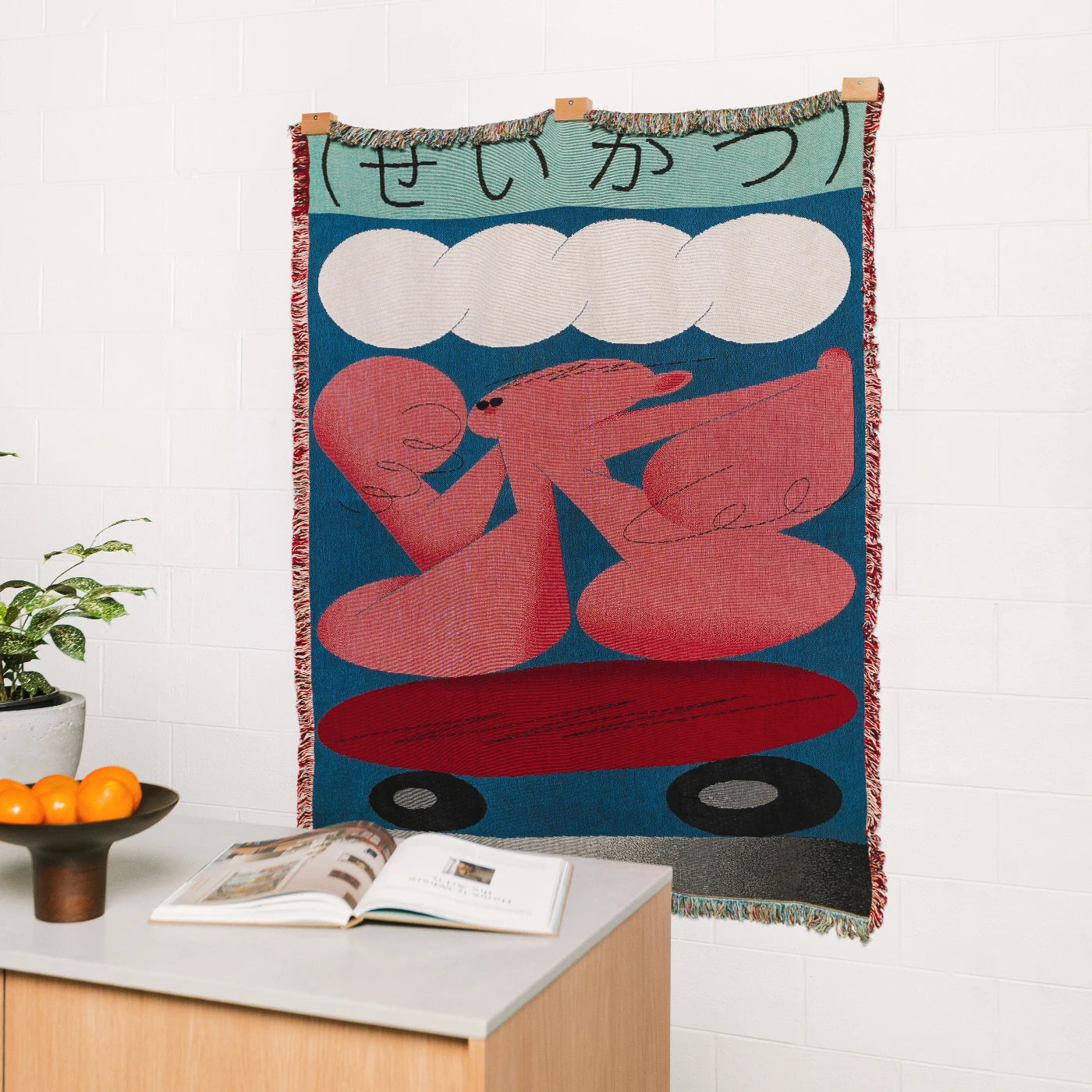 Decorative woven blanket with abstract design of a red figures on a blue background, with Japanese words, hanging on a wall above a table with Slowdown Studio blanket hanger clips. 