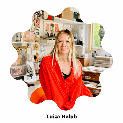 A portrait of Luiza Holub wearing a bright red draped top, seated in a cluttered creative studio. The photo is presented inside a decorative scalloped white frame with the text &