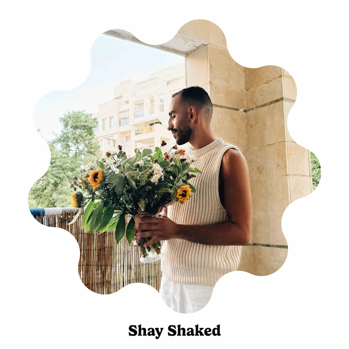 Artist Shay Shaked wearing a cream ribbed sleeveless sweater vest stands outdoors holding a large bouquet of sunflowers and greenery, framed by a decorative scalloped white border with the text &