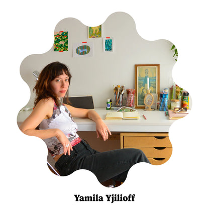 Argentinian artist Yamila Yjilioff sits at a white desk in a room with art supplies and framed wall art. 